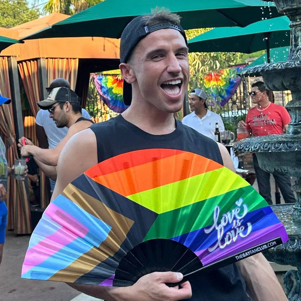 Hand fan featuring a vibrant Pride rainbow flag design from The Gay Fan Club, with colorful rainbow stripes and a fan handle.