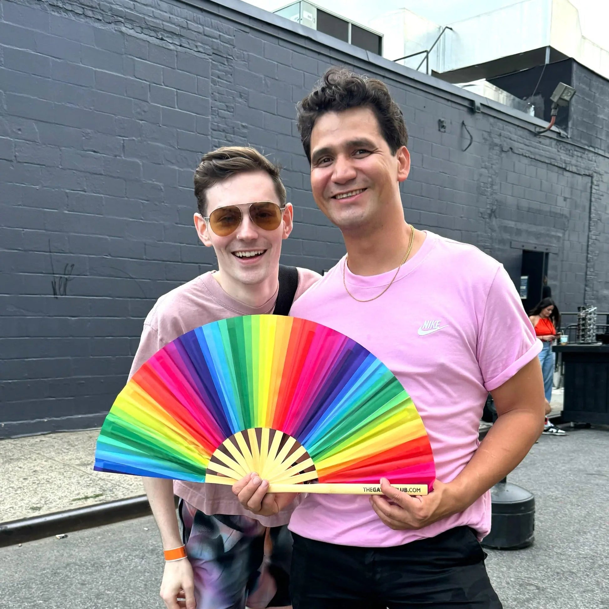 Rainbow folding hand fan by The Gay Fan Club featuring vibrant rainbow-colored fabric and a folding wooden handle.