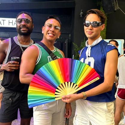 Rainbow folding hand fan by The Gay Fan Club featuring vibrant rainbow-colored fabric and a folding wooden handle.